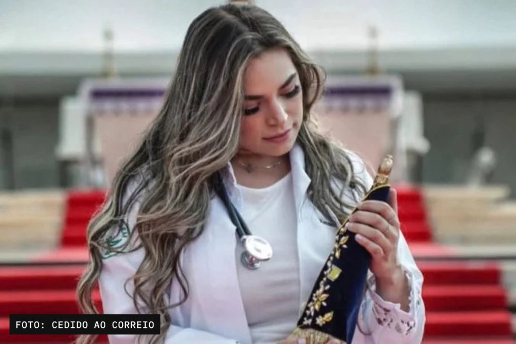 Foto de uma mulher adulta, identificável, usando jaleco branco e estetoscópio, segurando um objeto escuro decorado diante de uma escadaria.