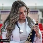 Foto de uma mulher adulta, identificável, usando jaleco branco e estetoscópio, segurando um objeto escuro decorado diante de uma escadaria.
