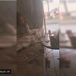 Foto vertical de um ambiente interno, com um sofá ou cama, uma pessoa deitada parcialmente desfocada à esquerda e uma mesa com bebidas e objetos.